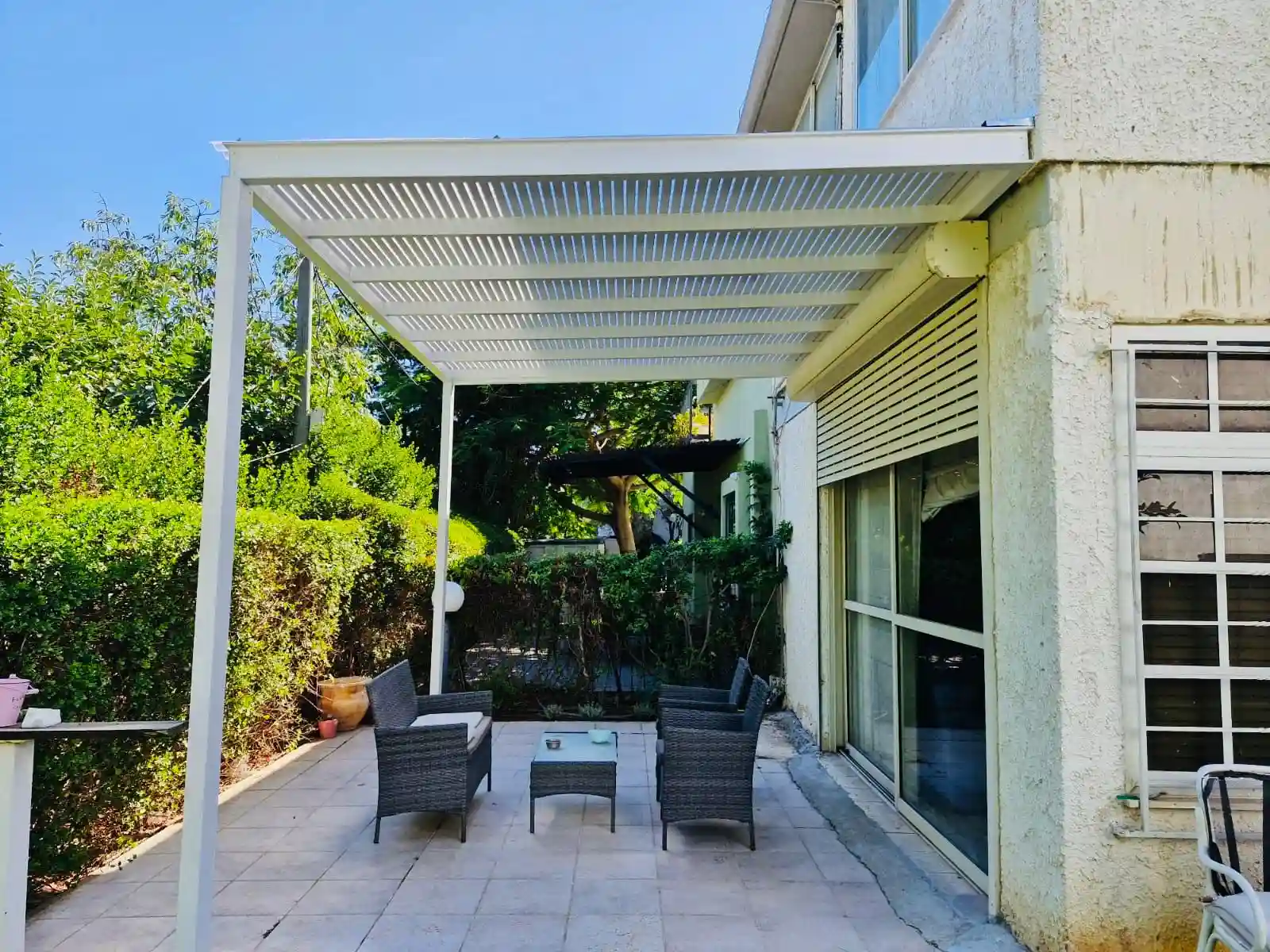 צילום חזיתי של פרגולה מאלומיניום בגוון לבן עם סרגלים צפופים מעל ריצוף גינה, מותאמת לפתח הסלון בבית פרטי