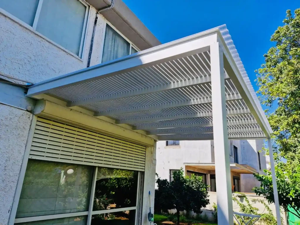 מבט אלכסוני על פרגולת אלומיניום לבנה עם סרגלים צפופים וסנטף BH, מותקנת על קיר חזית של בית דו-קומתי ברמת גן